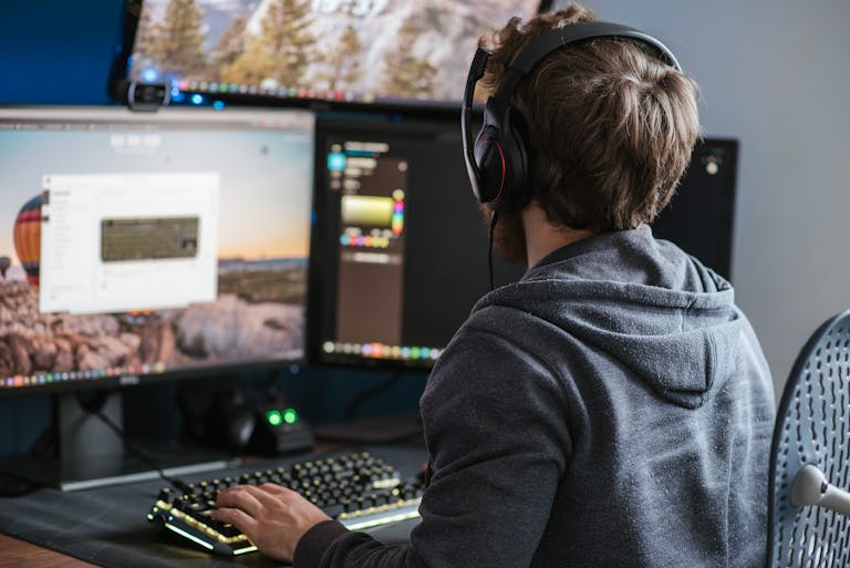 Adult man in casual attire working on dual monitors, wearing headphones, indoors.